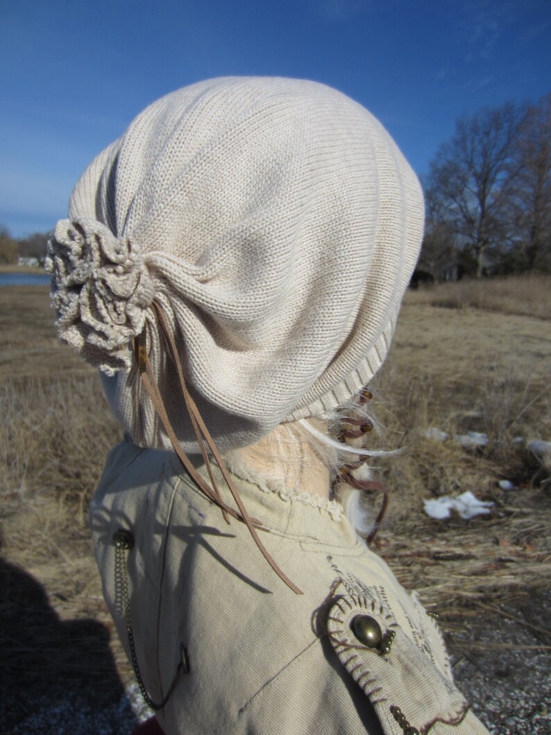 May include: A beige knitted beanie with a large flower detail on the side. The beanie has a long brown leather strap that hangs down the back.