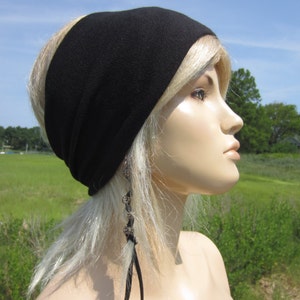 May include: A black headband made of soft fabric, worn by a person with blonde hair. The headband is wide and covers the forehead. The person is looking off to the side, with a field of green grass in the background.