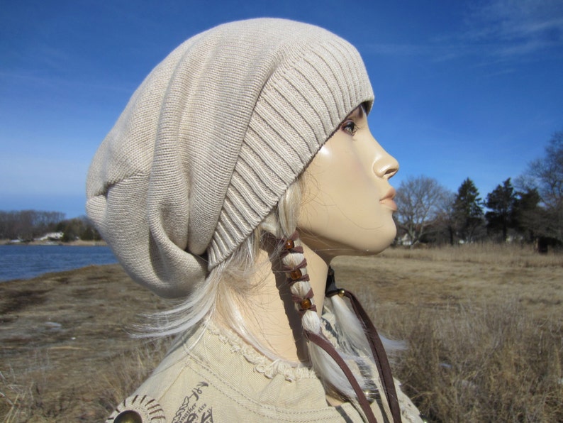 May include: A beige slouchy beanie hat with a long, white braid with brown beads hanging from the side.