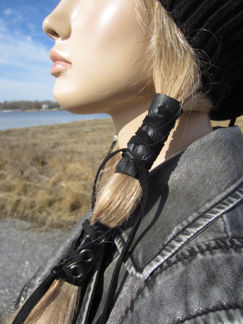 May include: A black leather hair tie with a braided design. The hair tie is wrapped around a ponytail of blonde hair.