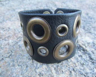 May include: Black leather bracelet with multiple large brass eyelets.