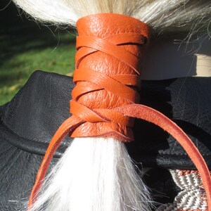May include: A bright orange leather hair tie with a braided design. The hair tie is tied around a ponytail of white hair.