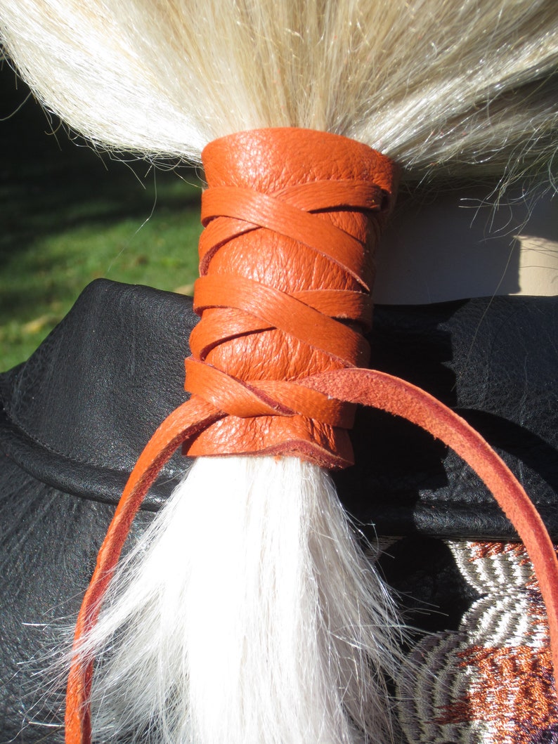 May include: A bright orange leather hair tie with a braided design. The hair tie is tied around a ponytail of white hair.