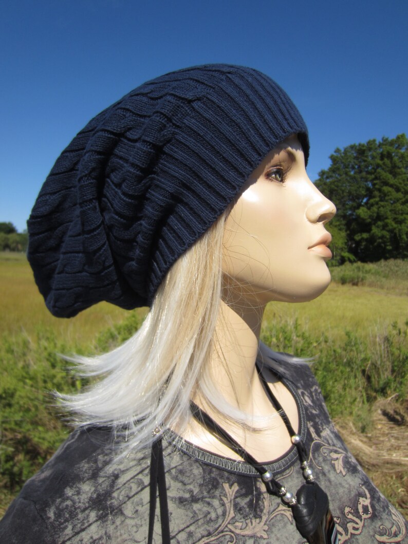 Puede incluir: Un gorro azul marino holgado con un patr&oacute;n de punto de cable. El gorro lo lleva una persona con el pelo largo y rubio y un collar negro con un colgante de plata.