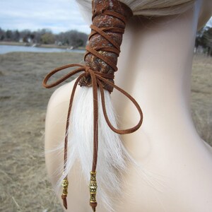May include: Brown leather hair tie with gold accents and a white faux fur tail. The hair tie is tied in a bow and has two gold beads on the ends of the tails.