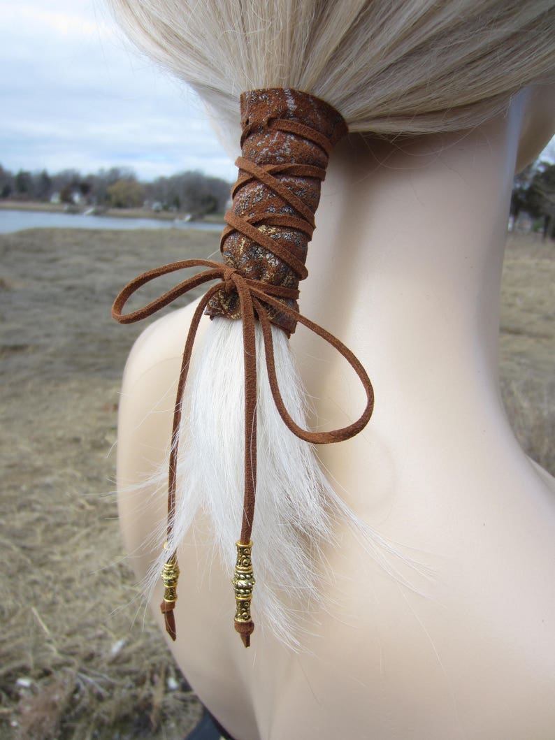 May include: Brown leather hair tie with gold accents and a white faux fur tail. The hair tie is tied in a bow and has two gold beads on the ends of the tails.