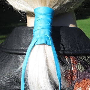 May include: A blue leather hair tie with long ties. The hair tie is wrapped around a ponytail of blonde hair.