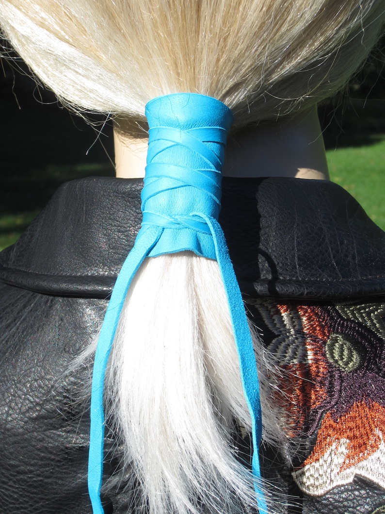 May include: A blue leather hair tie with long ties. The hair tie is wrapped around a ponytail of blonde hair.