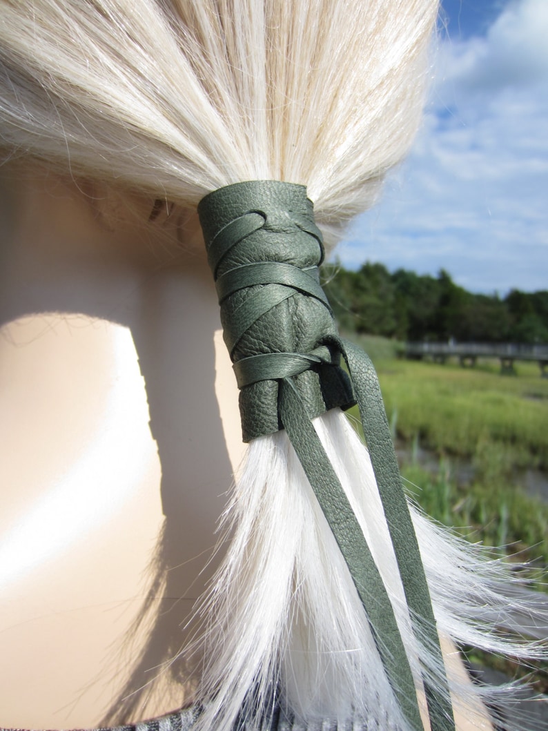 May include: A green leather hair tie with a braided design. The hair tie is tied around a ponytail of white hair.