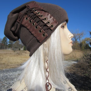 May include: A brown beanie with a leather lace-up detail. The beanie is worn on a mannequin with long white hair.