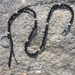 May include: Black braided lanyard with silver-colored accents and a metal clasp. The lanyard features small silver beads evenly spaced along the braid and a key ring. The lanyard is designed to hold keys or other small items.