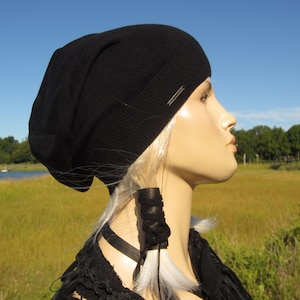 May include: A black beanie with a silver metal tag. The beanie is slouched and worn on a mannequin head with blonde hair. The mannequin is wearing a black top and has a black leather cord tied around its neck.