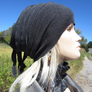 May include: A black slouchy beanie with a long tie. The beanie is made of a soft, knit fabric. The model is wearing a black leather jacket with a brown leather collar.