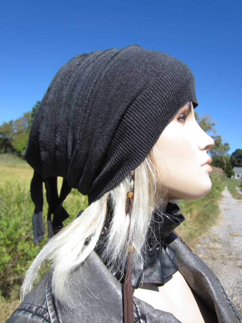 May include: A black slouchy beanie with a long tie. The beanie is made of a soft, knit fabric. The model is wearing a black leather jacket with a brown leather collar.