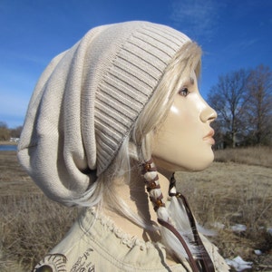 May include: A beige slouchy beanie with a long, flowing braid with brown beads. The braid is tied with a brown leather cord.