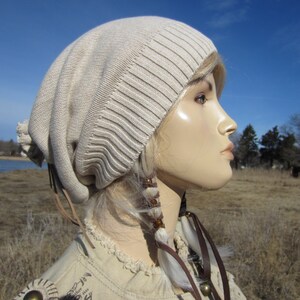 May include: A beige slouchy beanie with a brown leather strap and white feather accents. The beanie is worn on a mannequin with blonde hair.