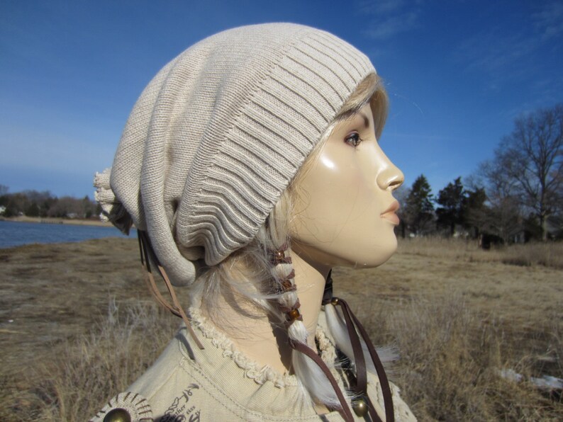 May include: A beige slouchy beanie with a brown leather strap and white feather accents. The beanie is worn on a mannequin with blonde hair.
