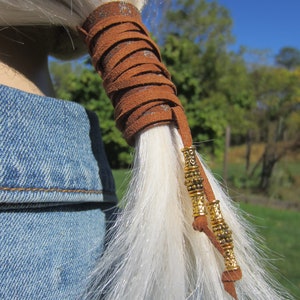May include: Brown leather hair tie with gold beads. The hair tie is wrapped around a ponytail of white hair.