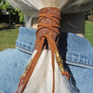 May include: A brown leather ponytail holder with gold beads and a decorative pattern. The holder is tied around a long, white ponytail.