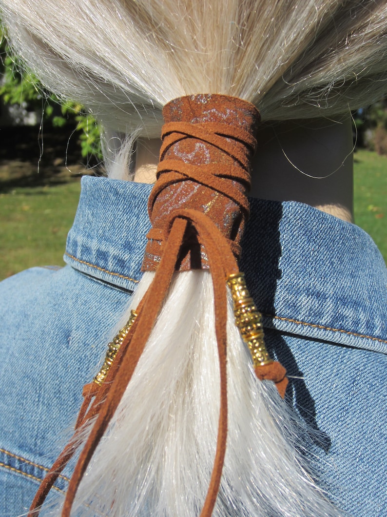 May include: A brown leather ponytail holder with gold beads and a decorative pattern. The holder is tied around a long, white ponytail.