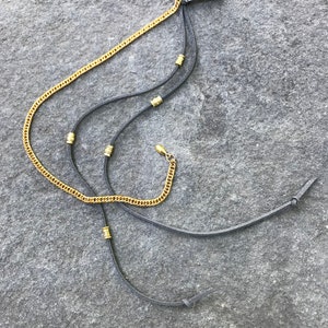 May include: A gold chain and two black leather cords with gold beads are attached to a black plastic hair clip.