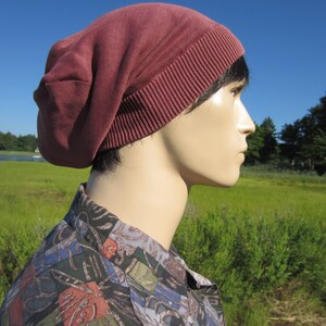 May include: A person wearing a maroon beanie with a folded brim. The beanie is made of a soft, knitted fabric. The person is standing in a field of green grass.
