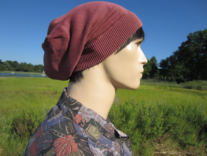 May include: A person wearing a maroon beanie with a folded brim. The beanie is made of a soft, knitted fabric. The person is standing in a field of green grass.