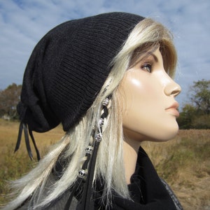 May include: A black beanie with a long black cord hanging down the side. The beanie is worn by a person with long, blonde hair. The hair is styled with a braid and silver beads.