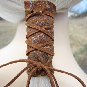 May include: Brown leather ponytail holder with a gold and silver patterned design. The holder is tied with a brown leather cord.