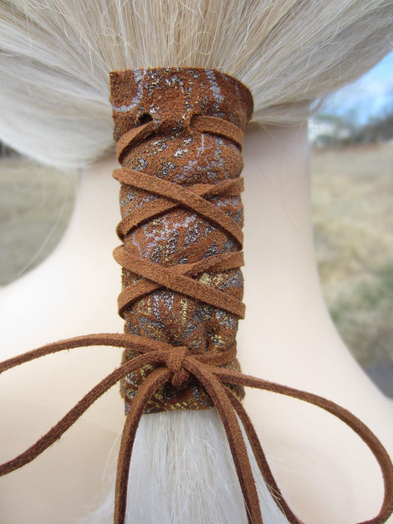 May include: Brown leather ponytail holder with a gold and silver patterned design. The holder is tied with a brown leather cord.