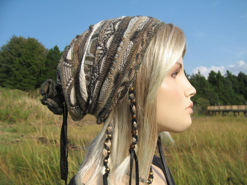 May include: A knitted beanie in shades of brown, beige, and black, featuring a textured pattern. The hat has a gathered design at the top and is paired with black ribbon ties and beaded accents. The background shows a grassy field and a blue sky.