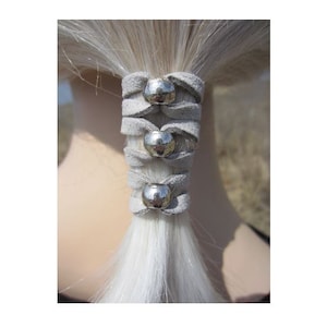 May include: A grey suede hair wrap, featuring three silver metal embellishments, is wrapped around a white ponytail. This hair accessory is designed to add a decorative touch to hairstyles, suitable for various occasions.