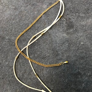 May include: A white leather cord with gold beads and a gold chain attached to a black hair clip.