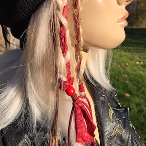 May include: Red and gold hair extensions with a red bead and tassels. The extensions are braided into blonde hair.