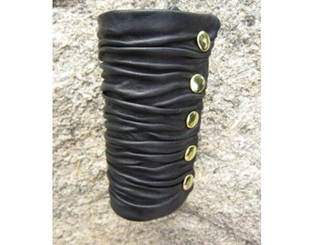 Op de afbeelding: Zwart leren armband met goudkleurige knopen. De armband is geplooid en heeft een textuur.
