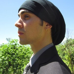 K&ouml;nnte beinhalten: Eine Person tr&auml;gt eine schwarze M&uuml;tze mit einem dunklen Anzugjackett und einer gemusterten Krawatte. Die M&uuml;tze ist aus weichem, dehnbarem Stoff und hat ein schlichtes Design.