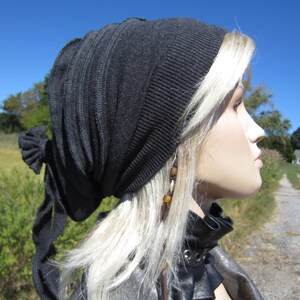 May include: A black knit beanie with a long, flowing tie. The beanie is worn on a person with long, blonde hair. The person is wearing a black leather jacket with a collar.