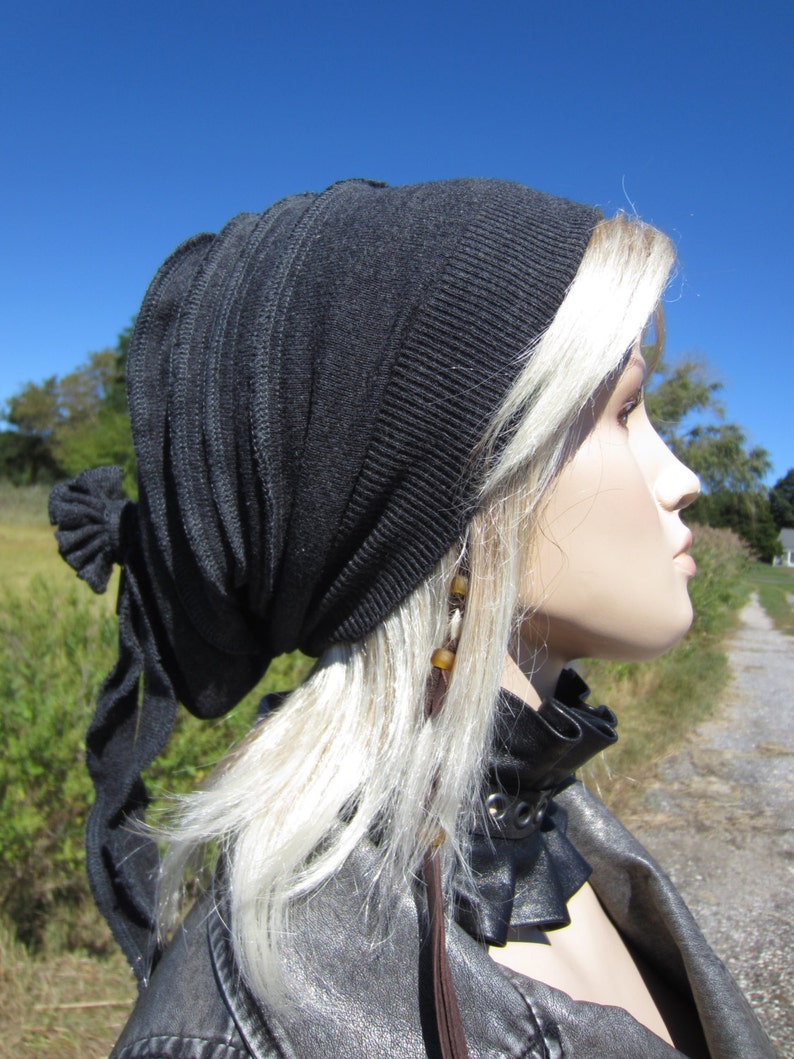 May include: A black knit beanie with a long, flowing tie. The beanie is worn on a person with long, blonde hair. The person is wearing a black leather jacket with a collar.