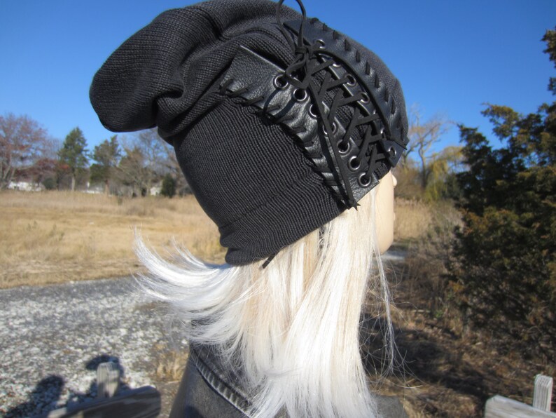 May include: A black beanie with a black leather lace-up detail on the side. The beanie is worn on a mannequin with long, white hair.