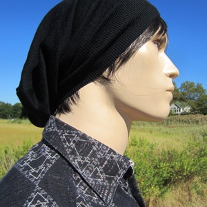 K&ouml;nnte beinhalten: Eine schwarze Beanie mit einem l&auml;ssigen Sitz. Die Beanie ist aus weichem, gestricktem Stoff gefertigt. Die Person, die die Beanie tr&auml;gt, tr&auml;gt auch ein schwarz-wei&szlig; gemustertes Hemd.