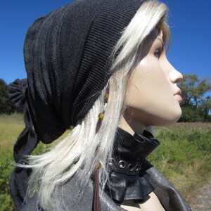 May include: A woman with blonde hair is wearing a black knit beanie with a long brown tassel. She is also wearing a black leather choker with silver grommets and a black leather jacket.
