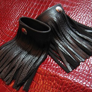K&ouml;nnte beinhalten: Zwei schwarze Lederarmb&auml;nder mit Fransen. Die Armb&auml;nder haben einen silbernen Knopfverschluss.
