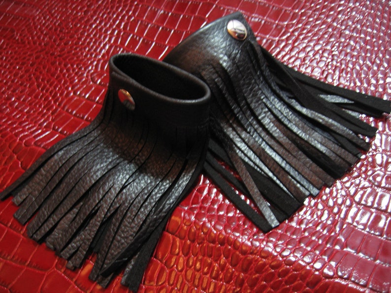 K&ouml;nnte beinhalten: Zwei schwarze Lederarmb&auml;nder mit Fransen. Die Armb&auml;nder haben einen silbernen Knopfverschluss.