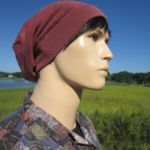 May include: A close-up of a person wearing a maroon beanie with a folded brim. The person is looking to the right and has short, dark hair. The background is a blurry image of a grassy field and a body of water.