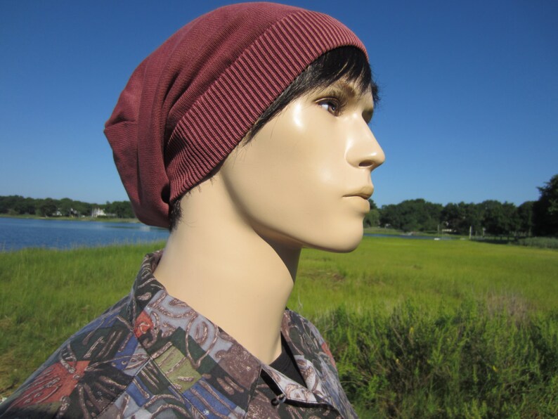 May include: A close-up of a person wearing a maroon beanie with a folded brim. The person is looking to the right and has short, dark hair. The background is a blurry image of a grassy field and a body of water.