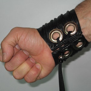 May include: Black leather wrist cuff with silver eyelets and a black cord tie. The cuff is worn on a person's wrist.