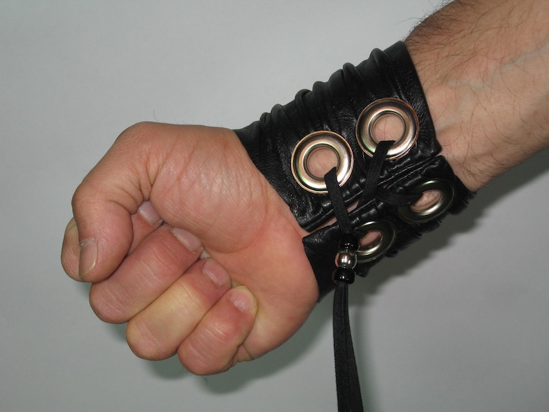 May include: Black leather wrist cuff with silver eyelets and a black cord tie. The cuff is worn on a person's wrist.