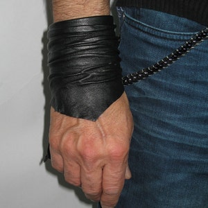 May include: A black leather cuff bracelet with a folded design. The bracelet is worn on the wrist of a person wearing blue jeans.