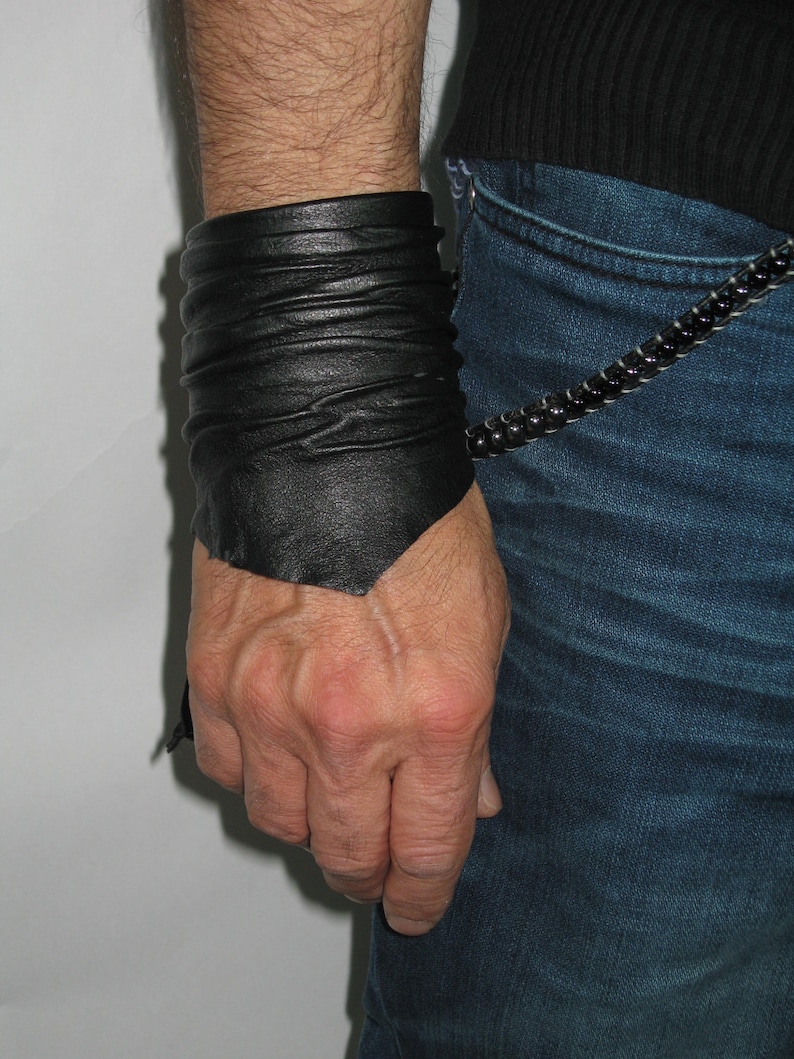 May include: A black leather cuff bracelet with a folded design. The bracelet is worn on the wrist of a person wearing blue jeans.