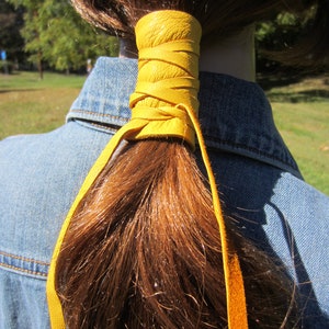 May include: A yellow leather ponytail holder with a braided design. The holder is tied around a brown ponytail.